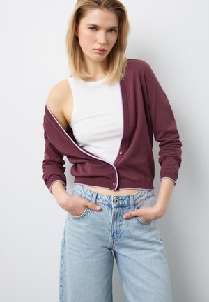 Giovane donna con canotta bianca, cardigan color amaranto parzialmente sceso sulla spalla e jeans a vita alta azzurri con le mani nelle tasche.