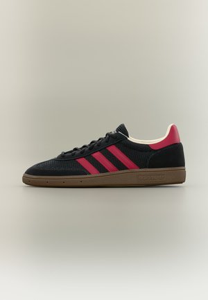 Sort Adidas sneaker med røde striber og hæl-accents, brun sål og sorte snørebånd, vist i sideprofil mod neutral baggrund.