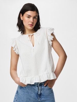 Frau mit dunklen Haaren trägt eine weiße, geraffte ärmellose Bluse mit Schleife am Hals und blaue Jeans, steht mit einer Hand in der Tasche.