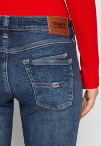 Jeans de mezclilla azul oscuro con una etiqueta de cuero marrón que dice "TOMMY JEANS". Presenta dos bolsillos traseros con una pequeña etiqueta de logo en rojo, blanco y azul.