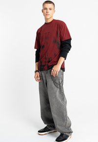 Czerwona i czarna koszulka tie-dye nałożona na czarną koszulę z długim rękawem, zestawiona z szerokimi szarymi dżinsami ozdobionymi łańcuchem. Całość dopełniają czarne trampki.