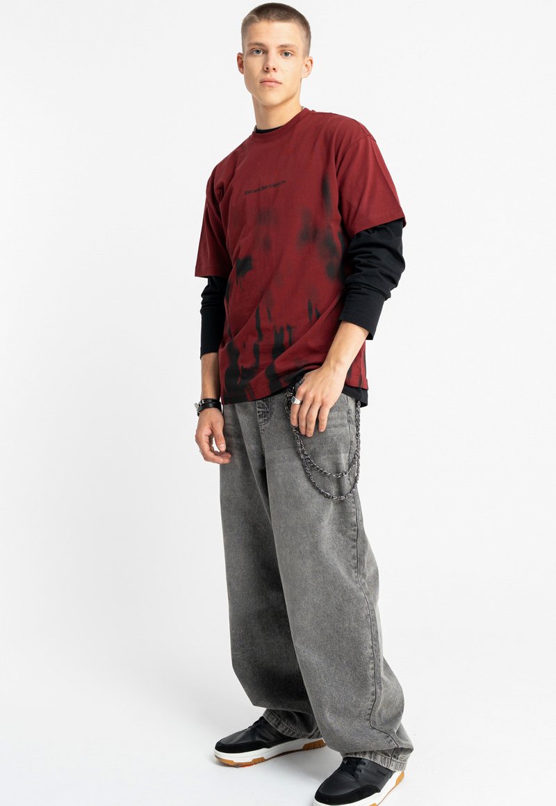 Czerwona i czarna koszulka tie-dye nałożona na czarną koszulę z długim rękawem, zestawiona z szerokimi szarymi dżinsami ozdobionymi łańcuchem. Całość dopełniają czarne trampki.