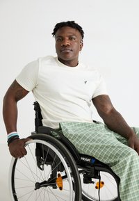 Fehér, testhezálló póló, zöld kockás nadrág és egy fekete kerekesszék narancssárga fényvisszaverőkkel. Az alany keze a kerék peremén pihen.