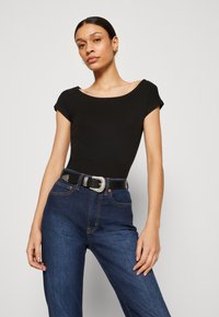 Top negru ajustat cu mâneci scurte; blugi închiși la culoare, cu talie înaltă; centură neagră cu detalii din metal argintiu. Țesătură netedă și croială structurată.