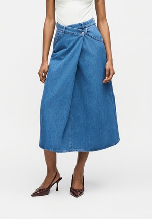 Modelka nosząca niebieską spódnicę dżinsową o długości midi z asymetrycznym zapięciem na guziki z przodu oraz brązowe szpilki slingback z zaostrzonym czubkiem.