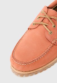 Zapato de barco de cuero coral con punta redondeada, cordones de color tierra, ojetes dorados y costuras visibles. Suela de goma texturizada y duradera con detalle de agarre.