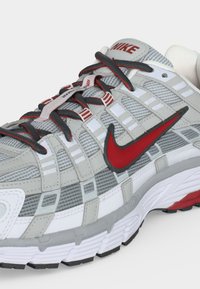 Ανδρικό γκρι και λευκό παπούτσι τρεξίματος Nike με κόκκινο λογότυπο swoosh και μαύρα-κόκκινα κορδόνια, που δείχνει λεπτομερή σχεδίαση διχτυού και σόλας.