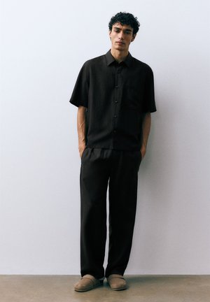 Homme aux cheveux bouclés portant une chemise noire ample à manches courtes avec boutons, un pantalon noir et des chaussons beige, debout contre un mur blanc uni.