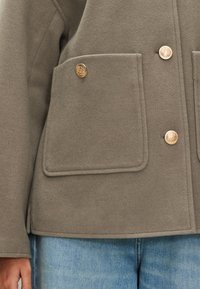 Pelēks fliša džemperis ar lielām priekšējām kabatām, zelta krāsas pogām un vaļēju piegriezumu. Tajā ir gluda tekstūra un minimālistisks dizains.