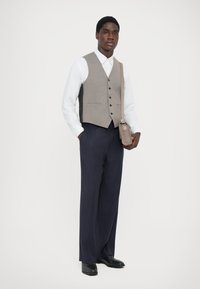 Grau Weste über einem weißen Hemd, kombiniert mit navy-blau gestreiften Hosen. Eine braune Umhängetasche und schwarze Schuhe runden das Outfit ab.