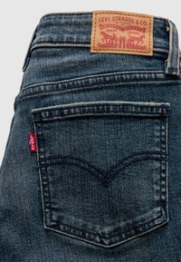 Jean en denim bleu avec une étiquette de marque en cuir, une couture arquée sur la poche arrière, et une petite languette rouge portant l'inscription "Levi's".