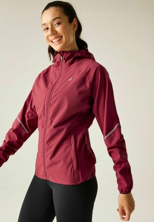 Woman smiling while wearing a maroon zip-up jacket with hood and reflective stripes on sleeves, standing against a plain background.