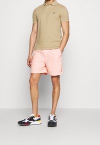 Polo beige con emblemma verde, pantaloni corti rosa chiaro e scarpe sportive multicolori con accenti bianchi e una fusione di texture.