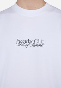 Fehér pamut póló, amelyen elegáns fekete kézírással a mellkasán a "Pegador Club" és a "Nyár Illata" felirat található.