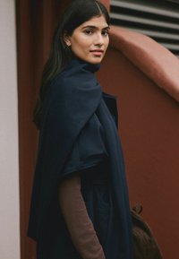 Woman with long dark hair wearing a navy blue cape coat and brown top, standing sideways near a curved reddish-brown wall and staircase.