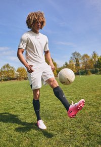 Fehér sportpóló és rövidnadrág, piros logóval, fekete térdzokni és rózsaszín focicipő, egy fűvel borított pályán focilabdát egyensúlyozva.