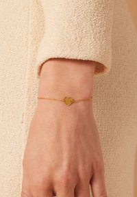 Gouden hartvormige armband op een delicate ketting, met een gladde oppervlakte en een minimalistisch ontwerp, tegen een gestructureerde beige stoffen achtergrond.