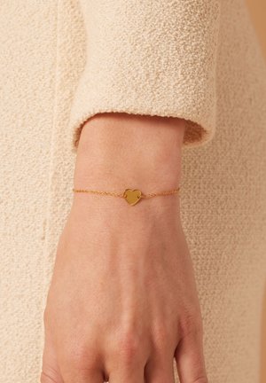 Bracelet en or en forme de cœur sur une chaîne délicate, avec une surface lisse et un design minimaliste, sur un fond en tissu beige texturé.