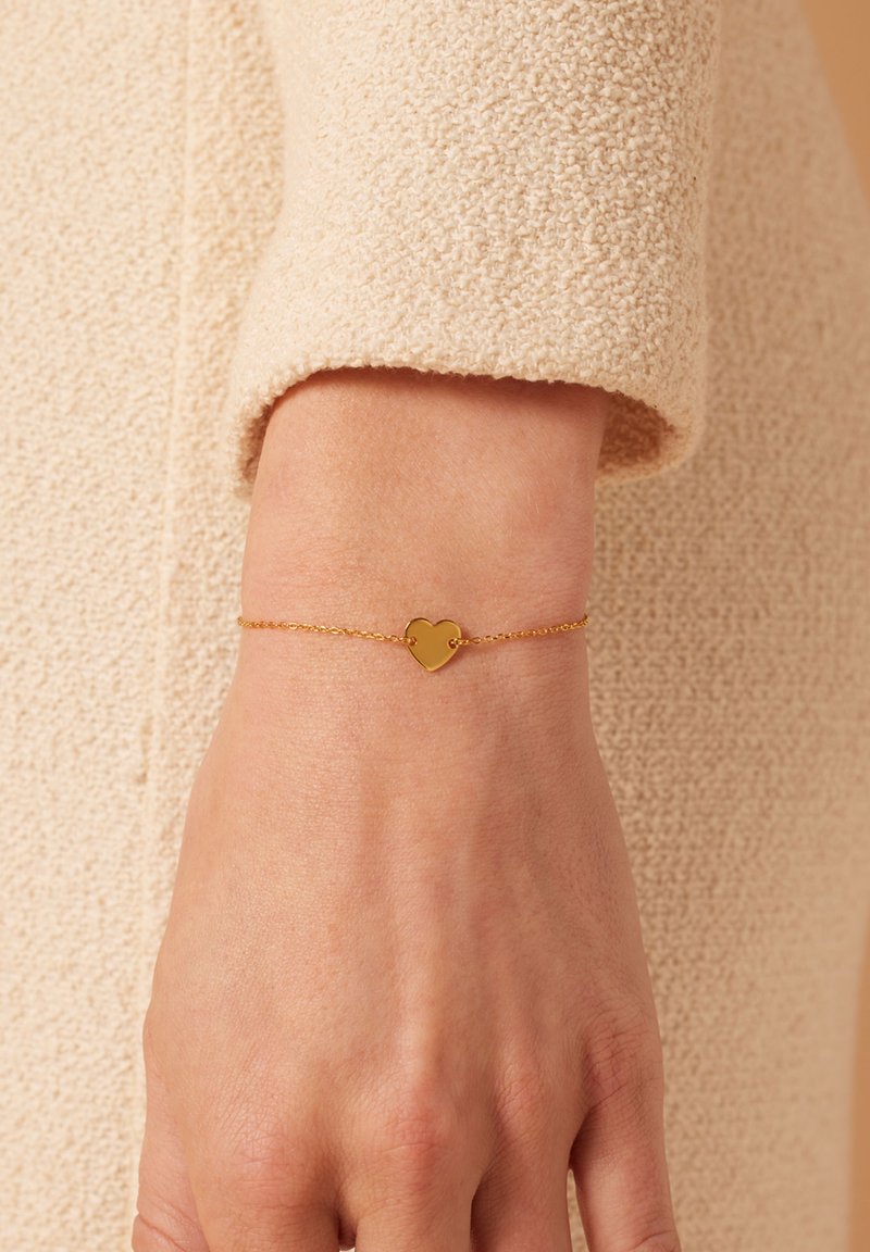 Gouden hartvormige armband op een delicate ketting, met een gladde oppervlakte en een minimalistisch ontwerp, tegen een gestructureerde beige stoffen achtergrond.