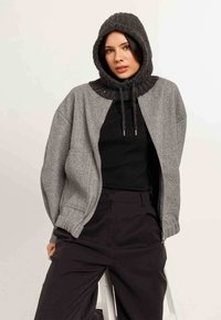 Giacca grigia con zip e cappuccio scuro testurizzato, colletto a coste e polsini elasticizzati. Indossata sopra una maglietta nera, abbinata a pantaloni scuri.