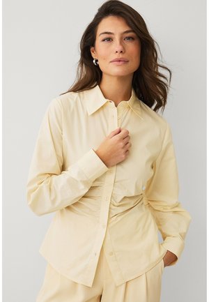 Kobieta z falującymi brązowymi włosami, nosząca srebrne kolczyki koła oraz jasnoszarą marszczoną koszulę zapinaną na guziki i pasujące spodnie, pozująca na jednolitym tle.