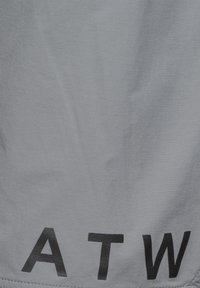 Grijze stof met grote zwarte letters "A T W" gedrukt vlak bij de onderrand, waarbij de textuur en lichte kreukels zichtbaar zijn.