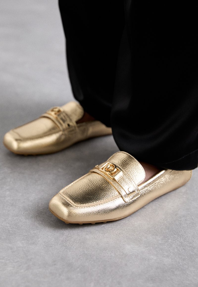 Gold loafers with a textured surface, featuring a decorative strap and emblem on the upper. Rubber sole for grip and comfort.