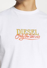 Bílý bavlněný tričko s nápisem "DIESEL" v tučné žluté barvě a "Only the Waves" ve vyšívané oranžové kurzivě. Krátké rukávy a design s kulatým výstřihem.