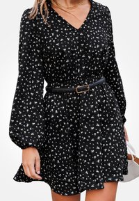 Donna che indossa un vestito floreale nero a maniche lunghe con scollatura a V e cintura nera, mentre tiene una borsa bianca con la mano destra.