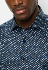 Blue patterned shirt featuring small white, blue, and brown designs, with a button-down collar and dark buttons. Texture appears smooth.