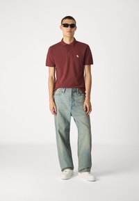 Burgunderrotes Poloshirt mit kleinem Logo, kombiniert mit hellblauen, baggy Jeans und weißen Sneakers. Das Modell trägt schwarze Sonnenbrille.