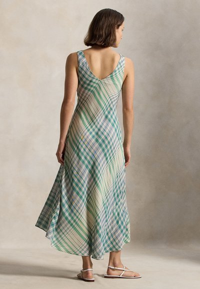 Γυναίκα που στέκεται με την πλάτη γυρισμένη, φορώντας αμάνικο μακρύ φόρεμα μέχρι τους αστραγάλους με καρό σχέδιο σε πράσινο, μπλε και λευκό, σε συνδυασμό με λευκά φλατ σανδάλια.