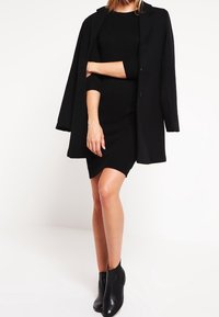 Abito nero a maglia con una silhouette aderente, abbinato a un lungo cappotto nero e stivali neri alla caviglia, caratterizzato da texture lisce e senza motivi visibili.