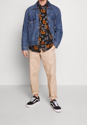 Mężczyzna w niebieskiej kurtce dżinsowej, czarnej koszuli w pomarańczowy kwiatowy wzór, beżowych spodniach z mankietami oraz czarnych trampkach z białymi sznurówkami i podeszwami.