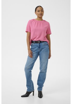 Femeie într-o bluză roz cu mâneci scurte, blugi albaștri cu curea neagră și ghete negre cu toc, stând pe un fundal alb simplu.