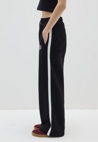 Pantalones de chándal negros con rayas laterales blancas, cinturilla elástica, bolsillos laterales y un detalle de logo. Llevados con zapatos tipo slip-on burdeos y beige.