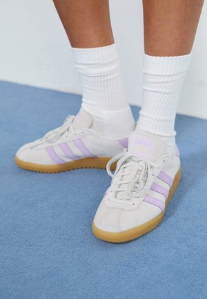 Fødder iført hvide sneakers med lyse lilla striber og gummisåler, parret med hvide ribstrikkede crew-sokker, står på blå tæppe.