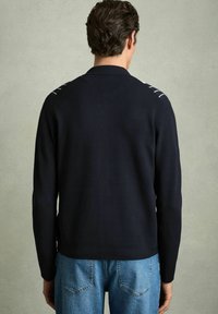 Pulover închis la culoare, de nuanță bleumarin, cu guler, având o textură cordată și linii albe accent pe umeri. Asortat cu blugi deschiși la culoare.