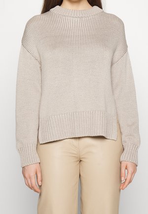 Femme portant un pull en grosse maille beige avec épaules tombantes et poignets côtelés, associé à un pantalon beige clair.