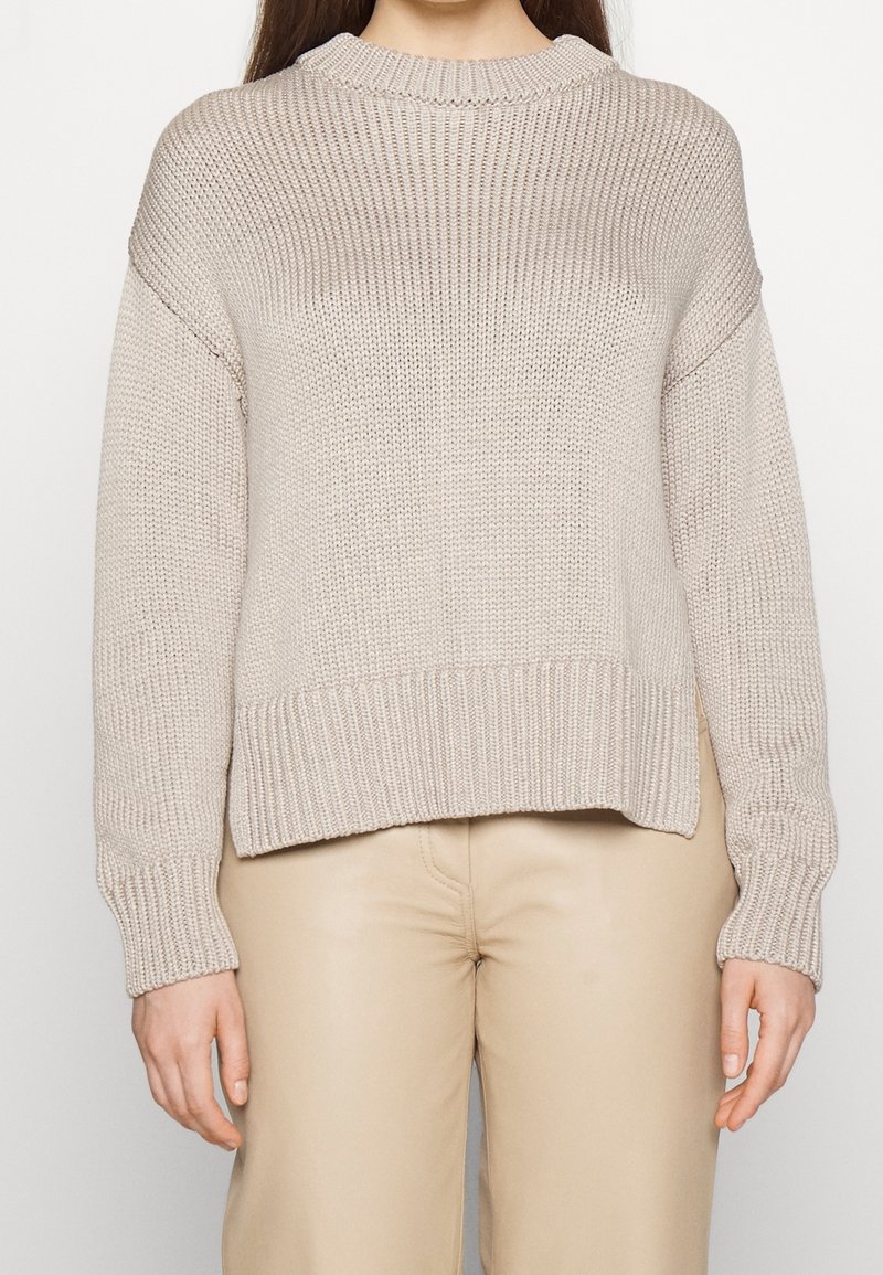 Femme portant un pull en grosse maille beige avec épaules tombantes et poignets côtelés, associé à un pantalon beige clair.