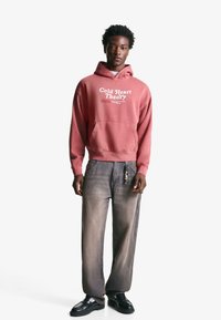 Hooded sweatshirt in faded pink featuring "Cold Heart Theory" graphic, paired with gray wide-leg jeans and black shoes. Smooth texture.