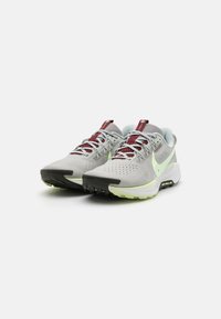 Un par de zapatillas de running Nike en gris y burdeos con suelas de un verde claro y punteras negras, inclinadas ligeramente hacia la izquierda sobre un fondo blanco.