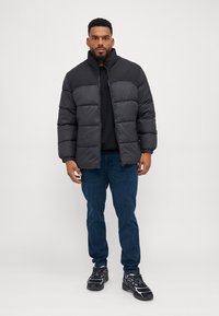 Sort pufferjakke med høj krave, firkantet form og quiltet tekstur. Båret over en sort sweater og parret med blå jeans og mørke sneakers.