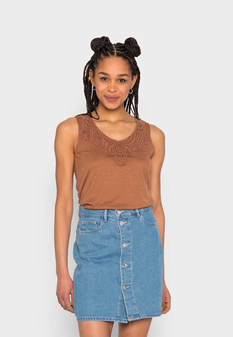 Top castanho sem mangas com decote bordado, combinado com uma saia de denim azul claro com botões à frente e bainha desfiada.