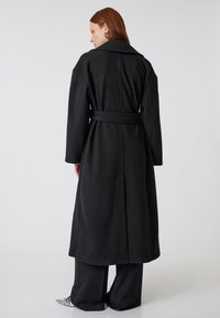 Zwarte wollen jas met een grote kraag, een ceintuur in de taille en lange mouwen. Bevat een lengte tot halverwege de kuit en minimale stikseldetails.