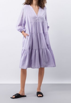 Femme portant une robe lilas ample à volants avec des manches trois-quarts et des sandales noires à enfiler, debout devant un fond uni.