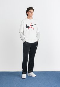 Balta grafika marškinėliai su juodu ir raudonu logotipu, uždėti ant pilkos ilgo rankovės marškinio. Juodos sportinės kelnės su mėlynomis šoninėmis juostelėmis ir baltomis sportinėmis bateliais.