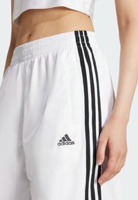 Biele teplákové nohavice vyrobené z ľahkého materiálu, s tromi čiernymi pruhmi po stranách a vyšívaným čiernym logom na boku.