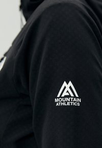 Manche de sweat-shirt noire avec logo blanc Mountain Athletics présentant des sommets de montagne stylisés au-dessus du nom de la marque.