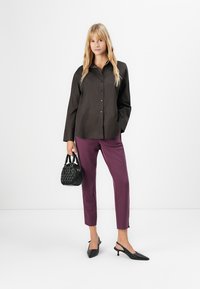 Ung kvinne med en svart skjorte med knapper, lilla capribukser, svarte høyhælte slingback-sandaler, som holder en liten svart håndveske.